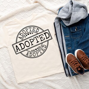 May include: A cream t-shirt featuring a black "ADOPTED" stamp design. A blue denim jacket with a grey hood and brown shoes are also visible. The items are arranged on a white, textured surface, suggesting a children's clothing theme.