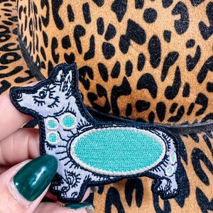 May include: A black, white, and teal corgi patch with intricate embroidery. The patch is held in front of a brown and black leopard print hat, creating a striking contrast. The patch features a teal oval design.