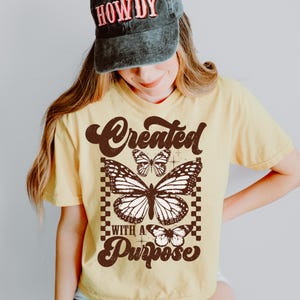 May include: Yellow t-shirt with a brown graphic of butterflies and the text "Created with a Purpose".