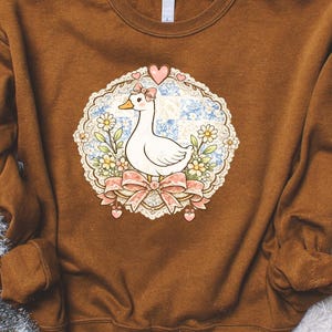 May include: A brown sweatshirt featuring a whimsical design of a white goose with a pink bow, set within a floral and heart-shaped frame. The goose is the focal point, surrounded by flowers and a decorative lace-like border.