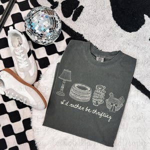 Op de afbeelding: Donkergrijs t-shirt met een witte afbeelding van een lamp, borden, kopjes en een mand met een kip. De tekst "I'd rather be thrifting" staat onder de afbeelding. Witte en grijze sneakers en een discobal staan ook in de afbeelding.