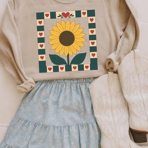May include: A beige sweatshirt with a sunflower design in a square frame with red hearts. The sweatshirt is paired with a light blue floral skirt and white cowboy boots.