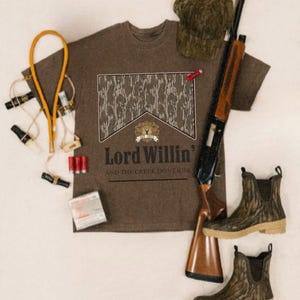 May include: A brown t-shirt with the text "Lord Willin'" and a camouflage design. The image also features a shotgun, a camouflage hat, boots, and hunting accessories.