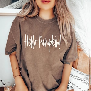 Może przedstawiać: Brązowy t-shirt z białym napisem "Hello Pumpkin!". Koszulka ma krótkie rękawy i luźny krój. Styl jest swobodny, odpowiedni na jesień. Tekst jest napisany odręcznie.