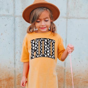 May include: A young child wearing a mustard yellow t-shirt with a black "MINI" graphic featuring a white ghost pattern. The child is wearing a brown fedora and holding a pink string.