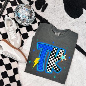 May include: A dark gray t-shirt with the letters "TK" in blue checkered and black and white checkered patterns, with a lightning bolt and stars. White and gray sneakers with black stripes and a disco ball are also in the image.