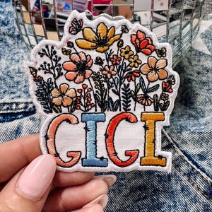 May include: An embroidered patch featuring a colorful floral arrangement with yellow, orange, and red flowers above the name GIGI in large, block-style letters. The letters are colored in peach, blue, red, and yellow with a white border.