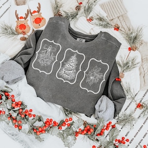 May include: A charcoal gray sweatshirt featuring three white framed holiday designs: mistletoe, a Christmas tree, and a poinsettia. The sweatshirt is folded with the sleeves tucked in. Decorative sprigs of greenery and red berries surround the sweatshirt.
