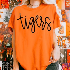 May include: Orange t-shirt with the word "tigers" in black script. The shirt is a solid orange color and has a crew neck. The text is in a cursive font.