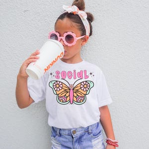 Może przedstawiać: Biała koszulka z nadrukiem motyla i tekstem "SOCIAL" w różowych literach. Motyl ma pomarańczowe, różowe i zielone skrzydła z białymi kwiatami.