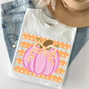 May include: A white t-shirt featuring a graphic of a pink pumpkin with a light orange bow, centered over horizontal orange stripes with white polka dots. The shirt is displayed alongside blue denim fabric and a tan wide-brimmed hat.