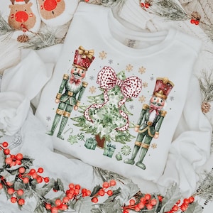 May include: White sweatshirt featuring a Christmas design with two nutcracker figures, a Christmas tree, and a large red bow. The design includes snowflakes, gifts, and candy canes. The sweatshirt is a Comfort Colors brand.