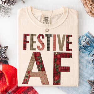 Puede incluir: Camiseta color crema con las palabras "FESTIVE AF" en un diseño de patchwork. Las letras están hechas de varios patrones, incluyendo cuadros y estampados florales. Se ve un collar plateado con un colgante de sol.