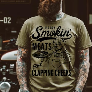 Könnte beinhalten: Ein grünes T-Shirt mit einer schwarzen Grafik eines Schweins, das einen Teller mit Fleisch hält. Der Text auf dem Shirt lautet "Old Row Smokin' Meats and Clapping Cheeks."