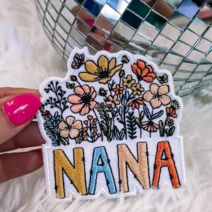 May include: An embroidered patch featuring a white background with a colorful floral arrangement at the top and the word NANA in large, multi-colored block letters at the bottom. The flowers are in shades of yellow, orange, pink, and blue.
