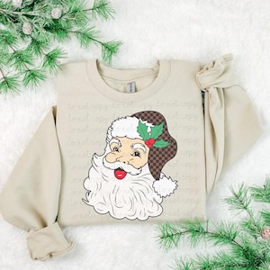 Puede incluir: Sudadera beige con un diseño de Papá Noel. El Papá Noel tiene barba blanca y un gorro a cuadros con detalles de acebo. Las mangas son largas y el estilo general es informal, ideal para la temporada navideña.