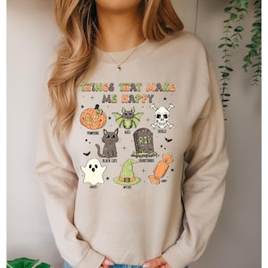 Könnte beinhalten: Beiges Sweatshirt mit einem farbenfrohen Halloween-Motiv. Das Design zeigt Kürbisse, Fledermäuse, Totenköpfe, schwarze Katzen, Geister, Hexen, Friedhöfe und Süßigkeiten, mit dem Text "Things That Make Me Happy".