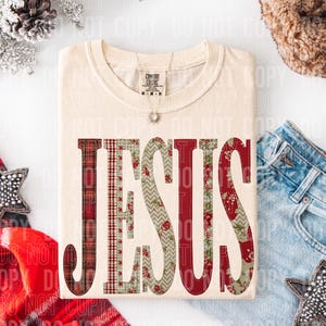 May include: A cream-coloured t-shirt featuring the word "JESUS" in a patchwork design of red plaid, floral, and chevron patterns. The shirt has a crew neck and short sleeves. A silver necklace with a sun pendant is visible.