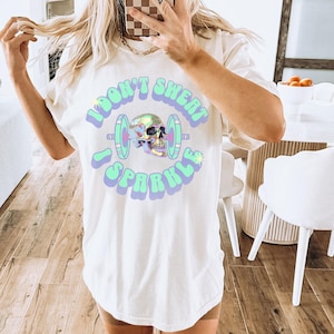 May include: White t-shirt with a graphic print of a skull holding a barbell. The text on the shirt reads "I don't sweat I sparkle".