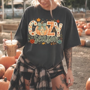 Puede incluir: Una camiseta gris carbón de manga corta con un estampado gráfico que incluye el texto "COZY season" en una fuente decorativa. El diseño presenta una calabaza central con un lazo verde azulado, rodeada de hojas de otoño, flores y cuadros.