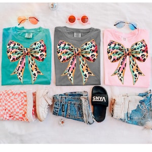 May include: Three t-shirts in teal, gray, and pink, each featuring a large bow design with a leopard print pattern in shades of pink, orange, and teal. Accessories include sunglasses, denim shorts, and a pair of Vans slides.