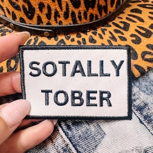 May include: A white rectangular patch with the words "SOTALLY TOBER" embroidered in black. The patch is held in front of a leopard print hat and denim fabric.