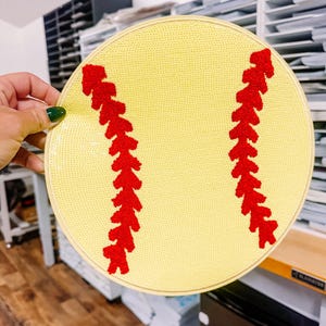 May include: A yellow softball embroidery hoop with red stitching detail. The hoop is round and has a light yellow background with red stitching to mimic a softball. The hoop is held by a hand with green nail polish.