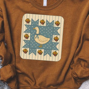 May include: A brown sweatshirt featuring a square graphic of a goose wearing a blue bow, centered on a blue and cream quilt-like design. The graphic is surrounded by yellow roses. The sweatshirt is a warm, earthy tone.