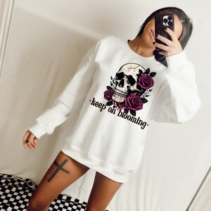 K&ouml;nnte beinhalten: Wei&szlig;er Pullover mit einem Totenkopf- und rosafarbenen Rosenmotiv und dem Text "keep on blooming".