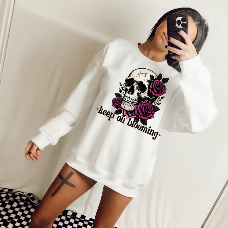 K&ouml;nnte beinhalten: Wei&szlig;er Pullover mit einem Totenkopf- und rosafarbenen Rosenmotiv und dem Text "keep on blooming".