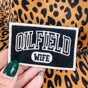 May include: A black and white patch with the words "OILFIELD WIFE" in a collegiate font. The patch is rectangular with a white border and is set against a leopard print background.