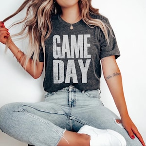 May include: Dark gray t-shirt with the words "GAME DAY" in large, distressed white letters. The shirt is a crew neck and appears to be made of a soft material. The overall style is casual and relaxed.