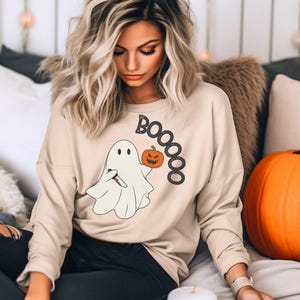 May include: A beige sweatshirt featuring a white ghost holding a jack-o'-lantern and the word "BOOOO" in black glitter.