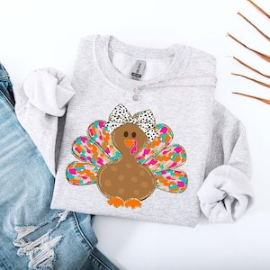 Könnte beinhalten: Ein graues Sweatshirt mit einem bunten Truthahn-Design. Der Truthahn hat einen braunen Körper mit weißen Punkten, einen rosa Schnabel und eine schwarz-weiße Schleife. Die Federn des Truthahns sind eine Mischung aus Rosa, Orange, Türkis und Gold.