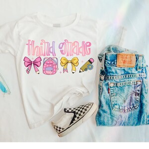 Pode incluir: Camiseta branca com o texto "Third Grade" em letras rosa e roxo. A camiseta tem um laço rosa, uma mochila de bolinhas rosa e roxo, um laço amarelo e um lápis amarelo com uma flor.
