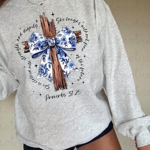 May include: A light gray sweatshirt featuring a wooden cross design with a blue floral bow. The text "She is clothed with strength and dignity" and "She laughs without fear of the future" surrounds the cross. The scripture "Proverbs 31:25" is also printed on the sweatshirt.