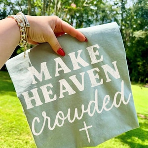 May include: A sage green t-shirt with the phrase "MAKE HEAVEN crowded" in white lettering and a white cross graphic. The shirt is held up against a background of green grass and trees.