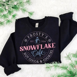 May include: Black sweatshirt with a circular graphic that reads "Frosty's Snowflake Cafe" in pink and blue. The design includes a snowman, snowflakes, and the text "Hot Cocoa & Cookies Served With Love."