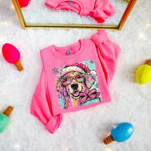 Puede incluir: Una sudadera rosa de cuello redondo con un gráfico colorido de un perro con gafas de sol y un gorro de Papá Noel. El perro sostiene un regalo con un lazo rosa. La sudadera está sobre una superficie blanca y esponjosa con luces decorativas.