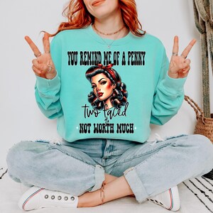 May include: A turquoise blue sweatshirt with the text "You remind me of a penny, two faced, not worth much" and a cartoon image of a woman with red lipstick and a red and white headband.