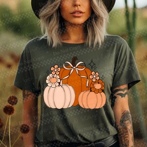 May include: Olive green t-shirt featuring a fall-themed graphic. The design includes a large orange pumpkin with a white bow, flanked by smaller pumpkins and floral accents. The shirt has a classic crew neck and short sleeves.