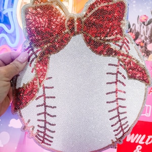 May include: A baseball-shaped decorative item with a large red sequined bow at the top. The baseball is white with red stitching and a silver sequined border. The bow has a gradient effect, transitioning from red to gold.