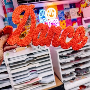 Pode incluir: Uma placa laranja e texturizada com a palavra "Dance" em itálico. A placa é segurada em frente a um fundo colorido com uma placa de néon e uma parede de várias imagens. A placa é um item decorativo.