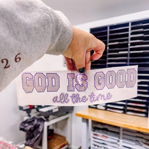 Könnte beinhalten: Ein weißes Band mit dem Schriftzug "GOD IS GOOD all the time" in lila Strasssteinen. Die Wörter sind fett gedruckt, mit dem Satz "all the time" in Kursivschrift. Das Band wird von einer Hand gehalten.