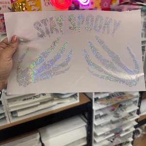 Può includere: Un foglio bianco con la scritta "STAY SPOOKY" e mani di scheletro, il tutto realizzato con strass iridescenti. Il design è probabilmente destinato a un progetto artigianale o alla decorazione di abbigliamento a tema Halloween.