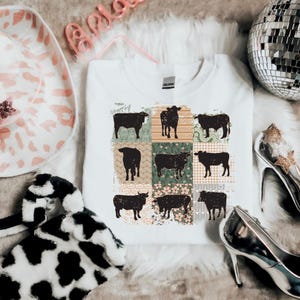 May include: White t-shirt with a patchwork design featuring black silhouettes of cows. The patchwork is made up of different floral and geometric patterns in shades of green, brown, and pink.