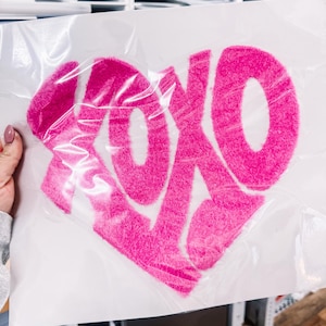 May include: A bright pink, heart-shaped design with the letters "XOXO" in a fluffy, textured font. The design is on a white background and is partially covered by clear plastic. The image is held by a person.