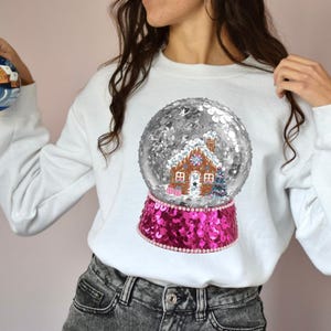 May include: White sweatshirt featuring a large snow globe design. The snow globe has a gingerbread house inside, surrounded by silver sequins. The base of the snow globe is pink and sequined.