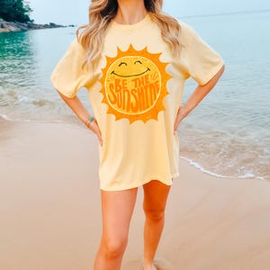 May include: A yellow t-shirt with a smiling sun graphic and the text "Be the Sunshine". The model is wearing the shirt on a beach.