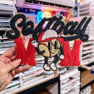 May include: A decorative sign with the words "Softball MOM" in black and red, with a baseball character. The letters are textured with gold trim. The baseball character is wearing a red hat and waving.